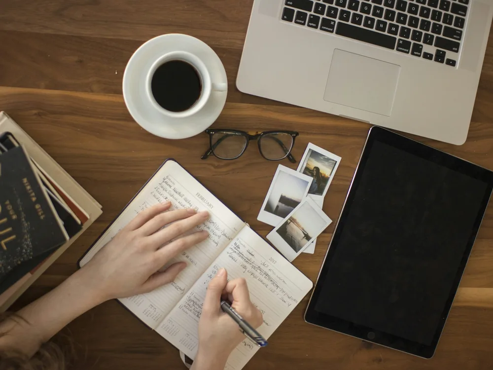 Hand writing in a notebook on a wooden desk beside a laptop, tablet and cup of coffee
