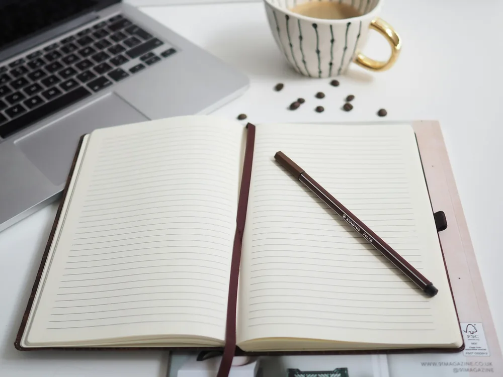 Open lined notebook and pen beside a laptop and a cup of coffee