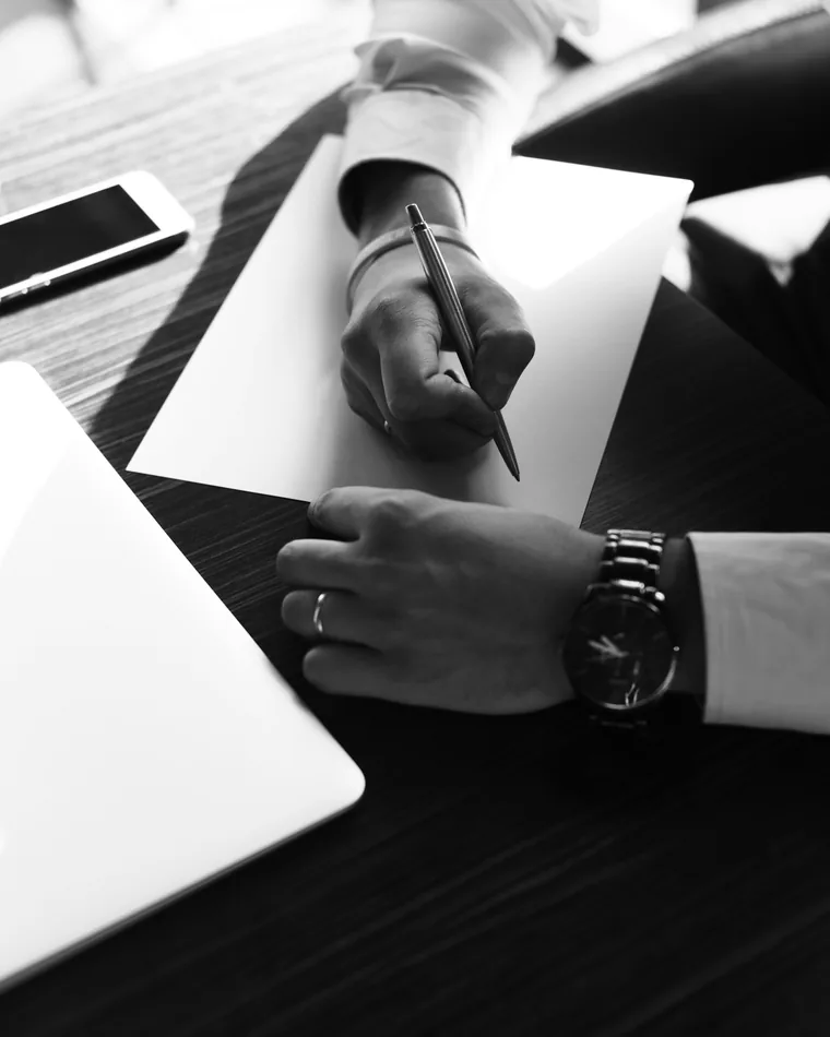 Black and white photo of a hand writing on paper beside a laptop