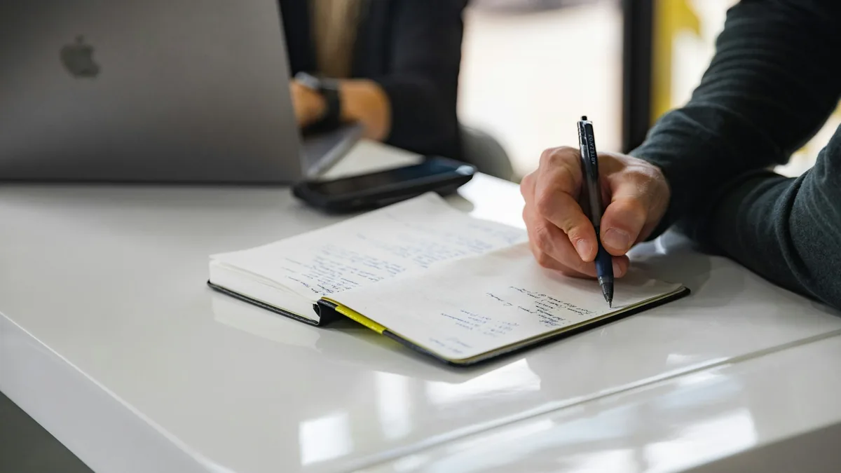 Hand writing notes in a notebook on a white desk next to a laptop
