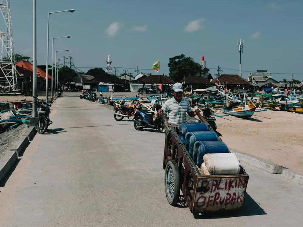 Man pushing a cart of water containers along a harbour road beside moored fishing boats